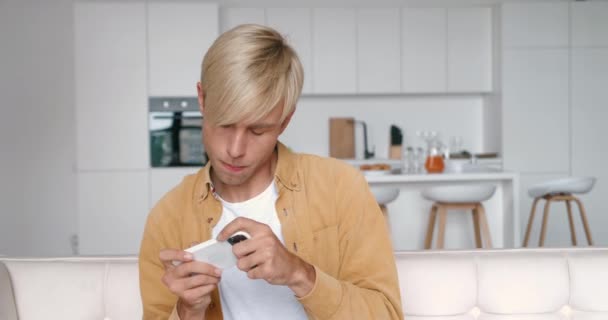 Jongeman spelen op zijn smartphone spannende mobiele applicatie winnen van een racespel knallende vuist vreugde terwijl zittend op de bank in de woonkamer thuis. — Stockvideo