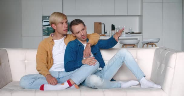 Glada män homosexuella par sitter hemma tillsammans på sin soffa i en omfamning. Manliga homosexuella par tillbringar tid hemma. HBTQ-förhållande — Stockvideo