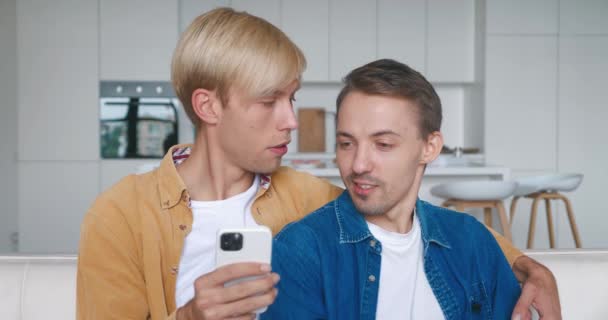 快乐的男同性恋夫妇一起坐在沙发上，用智能手机。男同性恋夫妇坐在客厅的沙发上拥抱和使用手机。LGBT关系 — 图库视频影像