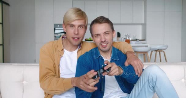 Emozionato divertente giovane maschio gay coppia gamers holding controller giocare video gioco insieme. Uomini LGBTQ felicissimi che hanno vinto il videogame divertendosi insieme sul divano celebrando la vittoria a casa. — Video Stock