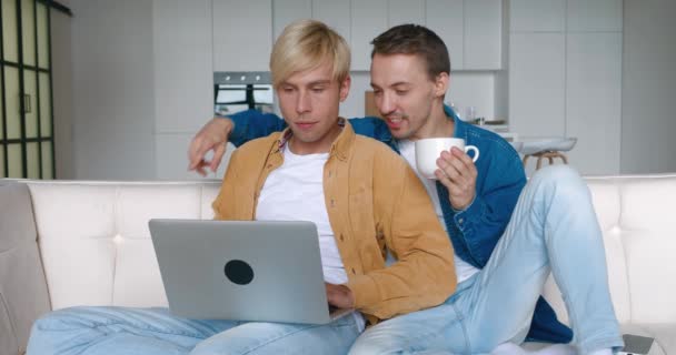 可爱的男同性恋夫妇会花时间呆在家里。两名LGBTQ男士在沙发上休息，并在客厅的沙发上使用笔记本电脑 — 图库视频影像