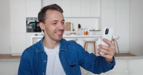 Jongeman in gesprek met smartphone camera bij het hebben van videocall binnen. Portret van een man met smartphone en videochatting en glimlachen terwijl hij thuis op de bank zit — Stockvideo