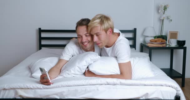 Homoseksuelle par kramme ser på smartphone kamera, når de foretager selfie fotos. To mænd bøsser liggende på sengen i soveværelset og tage billeder med mobiltelefon – Stock-video