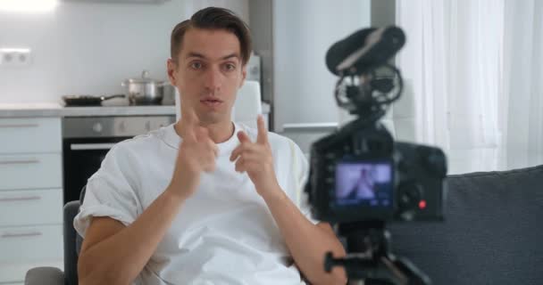 Блогер человек, снимающий новое видео видеоблога с профессиональной камерой, установленной на штатив дома, сидя на диване — стоковое видео