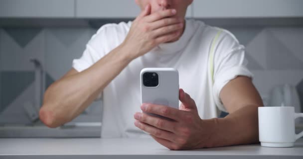 Close-up van een man surfen op het internet op een smartphone en is geschokt door het verschrikkelijke nieuws, bedekt zijn mond in angst terwijl thuis zitten in de keuken — Stockvideo