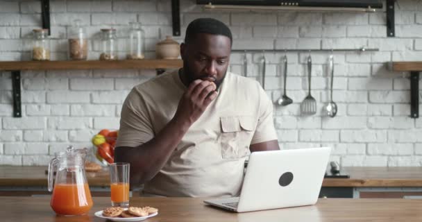 Afrikanischer Freiberufler isst Kekse und arbeitet am Computer, während er zu Hause am Küchentisch sitzt. — Stockvideo