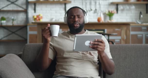 Sonriente hombre negro en los auriculares usando tableta digital sentarse en el sofá en casa, joven africano beber café sosteniendo la computadora mirando la pantalla de lectura aplicación de libro electrónico escuchar música en línea — Vídeos de Stock