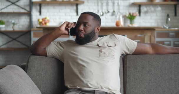 Hombre africano frustrándose mientras habla en su celular en su sala de estar en casa. Enojado hombre de negocios gritando durante la llamada telefónica sentado en el sofá en la sala de estar — Vídeos de Stock