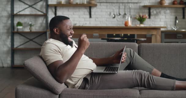 Счастливый молодой африканский бизнесмен празднует успех выиграть онлайн ставку и показать да жест. Счастливчик мужчина фрилансер чтение отличные новости в Интернете с помощью ноутбука во время отдыха на уютном диване дома. — стоковое видео
