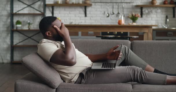 Trieste Afrikaanse zakenman werkt met laptop computer, verliest geduld terwijl hij thuis op de bank ligt. Ongeduldige mannelijke freelancer onder stress op het werk met pc in de woonkamer. — Stockvideo