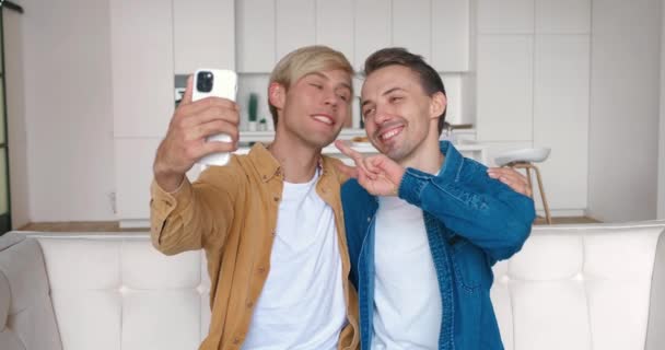 Гомосексуальная пара обнимается, глядя на камеру смартфона, когда делает селфи. Два гея фотографируют на мобильный телефон, сидя дома на диване. — стоковое видео