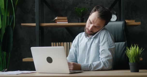 Multitasking Kaukasische jonge zakenman communiceren over zijn bedrijf op de mobiele telefoon tijdens het werken op laptop computer zittend op modern kantoor — Stockvideo