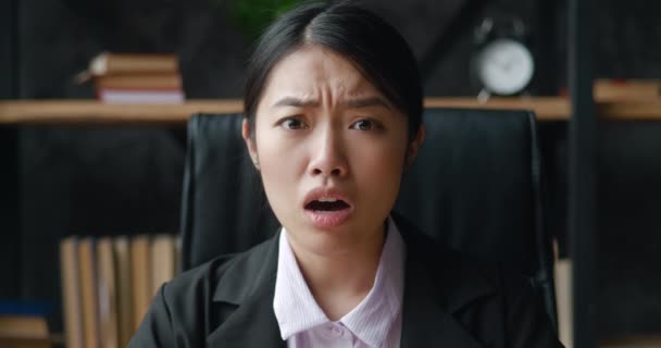 Chokeret asiatisk forretningskvinde ser på kameraet med frygt for forfærdelige nyheder, mens du sidder på kontoret. Portræt af bange kvindelige studerende, reaktion på dårlige nyheder, sidder indendørs på hjemmekontoret. – Stock-video