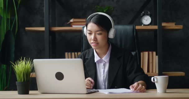 Asiatisk kvinna i hörlurar använder bärbar dator för distansundervisning och skriver anteckningar när du sitter på kontoret. — Stockvideo