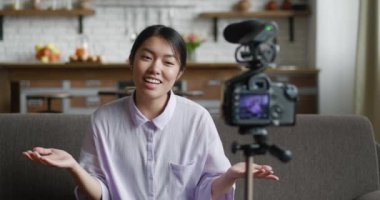 Asyalı kadın internet için video kaydediyor. Profesyonel video kamerayla tripoda monte edilmiş. Kadın blogcu evdeki koltukta oturuyor ve blog videosu kaydederken kamerayla konuşuyor