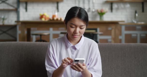 Atrakcyjna azjatycka kobieta siedząca na kanapie w domu przy użyciu telefonu komórkowego i uśmiechnięta. Komunikacja kobieta patrząc wiadomość na telefon komórkowy lub smartfona podczas siedzenia na kanapie w domu — Wideo stockowe