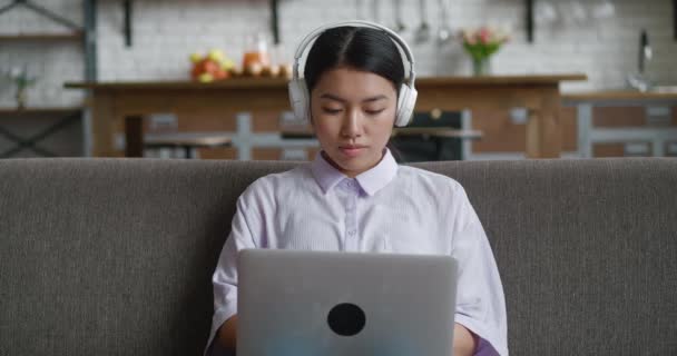 Asian woman with headphones resting on couch using laptop computer looking at screen typing message, listening music at home, young female browsing internet social media studying or working online — Stock Video