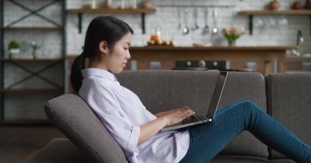 Mujer asiática descansando en el sofá usando portátil portátil mirando el mensaje de escritura de pantalla en la sala de estar, mujer joven charlando en la computadora navegando por Internet redes sociales estudiando o trabajando en línea en casa — Vídeo de stock