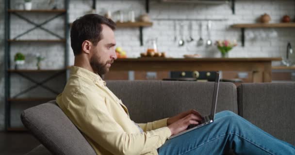 Hombre joven freelancer utilizando el dispositivo portátil mientras descansa en el sofá en casa, retrato de un hombre trabajando a distancia en el ordenador en el apartamento — Vídeo de stock