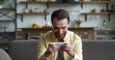 Akıllı telefondan oynayan genç adam heyecanlı mobil uygulama. Evdeki oturma odasında rahat bir kanepede otururken yumruğunu sıkarak bir yarış oyunu kazanıyor..