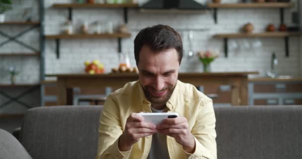 Fiatal férfi játszik okostelefon izgalmas mobil alkalmazás nyertes egy autóverseny játék szorító ököl örvendezik, miközben ül a kényelmes kanapén a nappaliban otthon. — Stock videók
