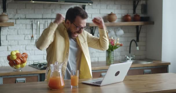 Ung affärsman sitter vid bordet med bärbar dator som arbetar i hemmakontoret kök bakgrund. Han får goda nyheter och är glad. Visar ja gest — Stockvideo