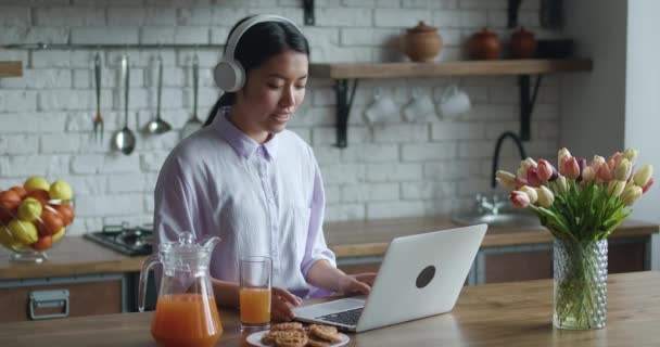 Junge fröhliche Asiatin sitzt am Küchentisch am Laptop und unterhält sich mit Freund online mit drahtlosen Kopfhörern aus ihren Ohren. — Stockvideo