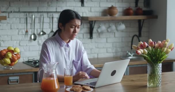 Frustrierte Asiatin, die zu Hause am Laptop arbeitet. Schockiertes besorgtes Mädchen, das enttäuscht auf den Computermonitor blickt, nachdem es eine Wette verloren hat, sitzt am Tisch in der Küche — Stockvideo