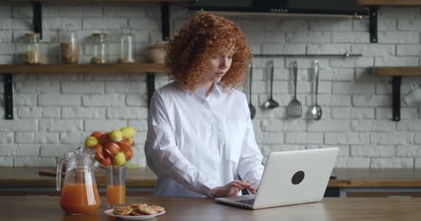 Redhead ung kvinna med lockigt hår sitta vid bordet i mysigt kök arbetar på bärbar dator, frilansare sms e-post svar till kunden, distansarbete aktivitet hemifrån med hjälp av internet och dricka juice — Stockvideo