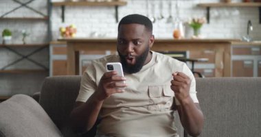 Şaşırtıcı Afrikalı erkek kazananın akıllı telefonu var. Evdeki mobil online bahis oyununun kazanmasından hayrete düşmüş iyi haberler okuyor. Mutlu erkek, koltukta otururken zaferin verdiği sevinçle cep telefonu ekranına bakar..