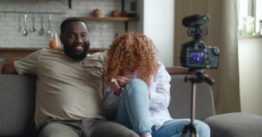 İnternet için video kaydeden blogcular tripoda monte edilmiş profesyonel video kamerayla evdeki koltukta oturuyorlar. Afrikalı erkek ve beyaz kadın kamerayla konuşuyor, oturma odasında blog kaydediyor.