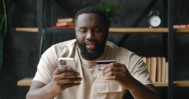 Mutlu Afrikalı Amerikalı iş adamı elinde kredi kartı ve cep telefonu ile internet üzerinden alışveriş yapıyor, dilek tutulan şeyleri alıyor, evet diye bağırıyor, ev konseptinden alışveriş yapıyor.