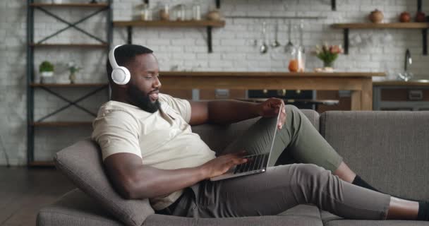 Vista lateral hombre afroamericano en auriculares apoyados en el sofá, trabajando de forma remota en el ordenador portátil desde casa. Empresario charlando en la computadora navegando por internet redes sociales estudiando o trabajando en línea — Vídeos de Stock