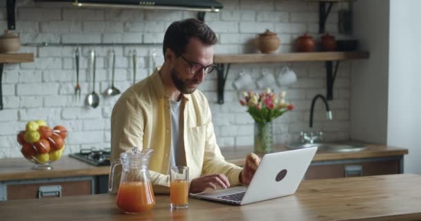 Junger Mann mit Brille, der am Laptop arbeitet und zu Hause in der Küche leckeres Croissant mit Saft isst. Kaukasischer Geschäftsmann, Tippnachricht, Internetsurfen in der Küche. — Stockvideo