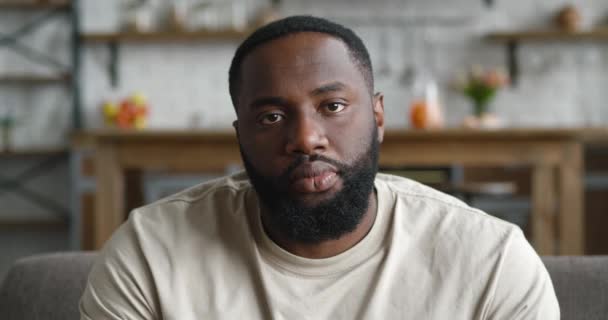 Retrato de cerca del guapo joven afroamericano barbudo, sentado en el sofá en casa en la sala de estar, usando camiseta, mirando a la cámara — Vídeo de stock