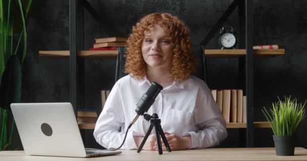笑顔のインフルエンサー赤髪の女性は、プロのマイクロフォンでキャストビデオを記録します.デジタルブロガーvloggerストリーミングトークショーホームオフィススタジオで、デスクに座って. — ストック動画