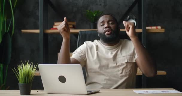 Empresário africano feliz dançando alegremente enquanto trabalhava em laptop no escritório em casa. Trabalhador masculino engraçado sentado na mesa, trabalhando no computador on-line, lendo notícias positivas, comemorando, dançando com alegria — Vídeo de Stock