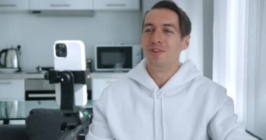 Mutlu blogcu adam evde akıllı telefon kamerasıyla video kaydediyor. Erkek kanepede oturuyor ve video konferansı veya video görüşmesi yapıyor. Vlog içeriği oluşturucu cep telefonuyla vlog kaydediyor