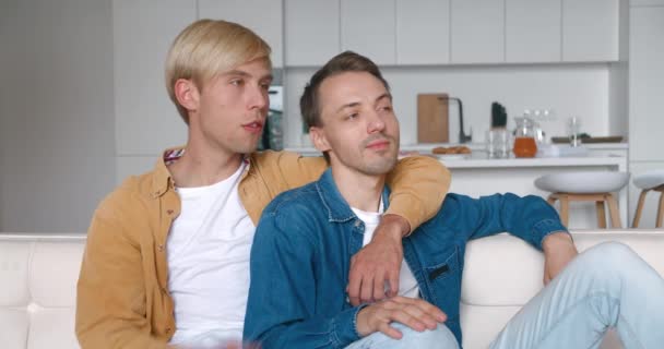 Ευτυχισμένοι άντρες ομοφυλόφιλοι κάθονται στο σπίτι μαζί στον καναπέ τους σε μια αγκαλιά. Αρσενικό γκέι ζευγάρι περνάει χρόνο στο σπίτι. Σχέση ΛΟΑΤ — Αρχείο Βίντεο