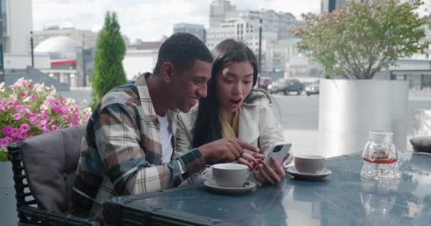 Charmant jong stel in casual gezellige kleren zitten aan de tafel in een zomerterras van een cafetaria op zonnige warme vroege voorjaarsdag en kijken naar een smartphone, verheugen en knuffelen elkaar. — Stockvideo