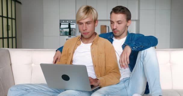 Шокированная гей-пара смотрит на экран ноутбука и расстроена плохими новостями, сидя вместе на диване дома — стоковое видео