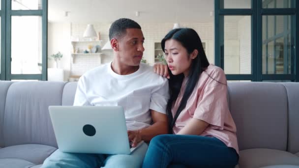 Pasangan muda antar ras duduk di sofa di rumah dengan laptop dan situs selancar, membahas bersama-sama, penuh perhatian menonton film. — Stok Video