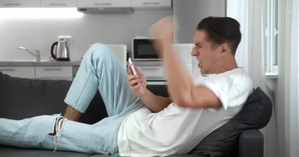 Genervter junger Mann, der sein Smartphone frustriert in der Hand hält und sich zu Hause auf dem Sofa ausruht. Wütender Mann benutzt Anwendung auf seinem Handy, schlägt mit der Faust auf das Sofa und schreit. Aggressivität zur Schau gestellt. — Stockvideo