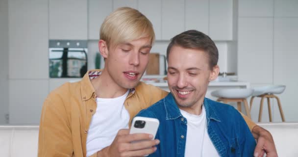 Dois homens casal homossexual sentar em casa juntos em um abraço em seu sofá e usando smartphone. Casal gay masculino passa tempo em casa. Relacionamento LGBT — Vídeo de Stock