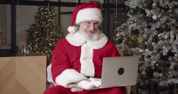 Όμορφος ώριμος άντρας με στολή καρναβαλιού του Άη Βασίλη που αναπαύεται στο σαλόνι κοντά στο χριστουγεννιάτικο δέντρο και πληκτρολογεί στο πληκτρολόγιο του φορητού υπολογιστή, διαβάζοντας email με τις επιθυμίες των παιδιών — Αρχείο Βίντεο