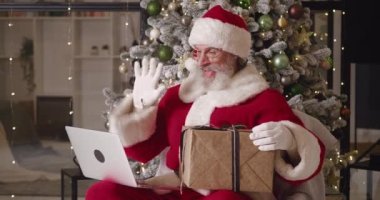Neşeli Noel Baba koltuğunda otururken evinin arka planında bir Noel ağacına karşı, video görüşmesi sırasında sohbet ediyor, baş parmağını gösteriyor ve bir Noel hediyesi kutusu yolluyor.