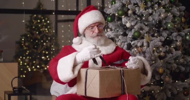 Przystojny Święty Mikołaj koncentrując się na pakowaniu pudełka z prezentami, próbując zawiązać kokardę satynową wstążką, a następnie radując się po zakończeniu, uśmiechając się radosnym, zębowym uśmiechem patrząc w obiektyw — Wideo stockowe