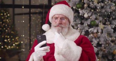 Düşünceli Noel Baba evinde süslü Noel ağacına karşı duruyor, akıllı telefondan çocukların mesajlarını okuyor, etrafa dikkatlice bakıyor ve başını güzel bir gülümsemeye doğru sallıyor.