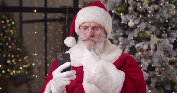 O gentil e pensativo Papai Noel está contra a árvore de Natal decorada em sua residência, lê mensagens de crianças em um smartphone, olha atentamente ao redor e acena com a cabeça espalhando-se em um belo sorriso — Vídeo de Stock