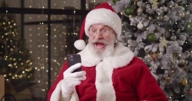 Neşeli Noel Baba, akıllı telefonunda mesajlar okuyarak, gülümseyerek, gülerek, başparmağını kaldırarak, mutluluktan yumruklarını sıkarak çocukların Noel dileklerini yerine getirebilmek için sevinir.